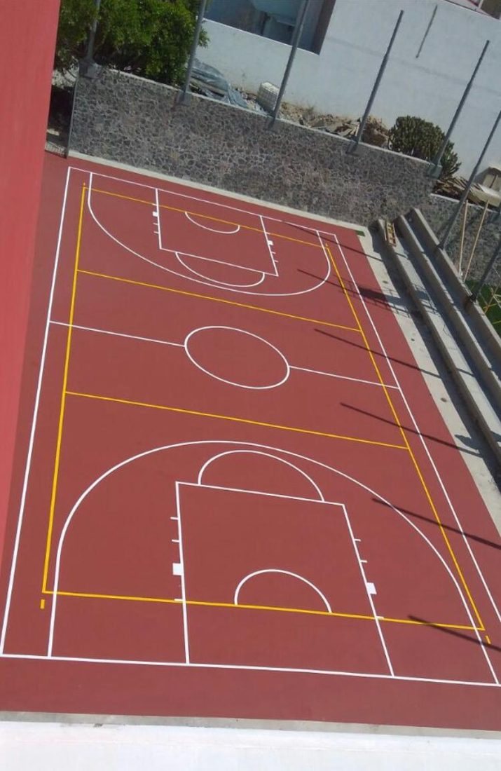 Cancha de básquetbol con superficie acrílica roja y líneas blancas y amarillas, junto a un muro de piedra