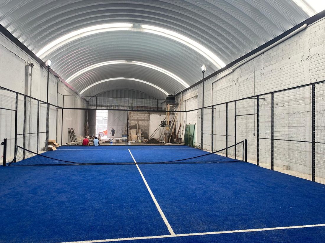Cancha de pádel de pasto azul dentro de una nave con arcotecho de lámina y muros de block