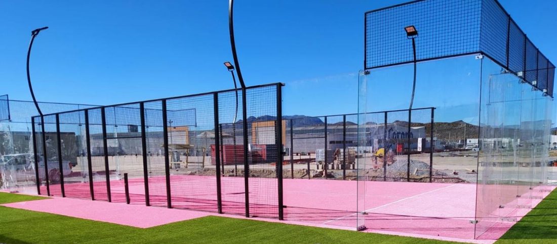 Cancha de pádel con piso rosa y paredes de cristal al aire libre, rodeada de pasto