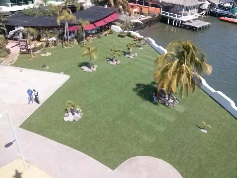 Jardín de pasto sintético con palmeras junto al agua y un muelle, visto desde arriba